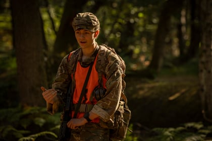 A person in military camouflage and an orange vest gestures with their left hand whilst standing in a sunlit forest.
