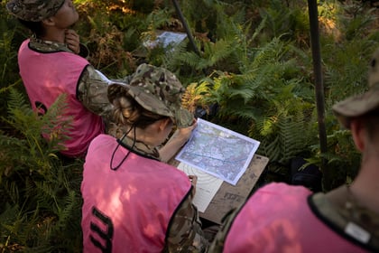 Three people in military uniforms and pink vests study a map on a table in a forested area with dense green ferns, preparing or planning an activity.