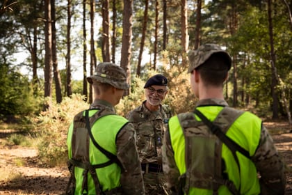 Three soldiers in camouflage uniforms and neon vests stand in a sunlit forest, facing an older officer who is smiling and speaking to them. Tall trees and greenery are visible in the background.