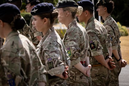 A group of people in camouflage military uniforms stand in a line outdoors, facing forwards with their hands clasped behind their backs.