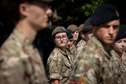 A group of young people in camouflage uniforms stands in formation outdoors; one person with glasses and a beret looks directly at the camera whilst others face forwards. Trees are blurred in the background.