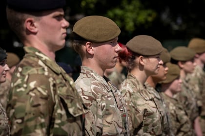 A group of soldiers in camouflage uniforms and berets stand in formation outdoors, facing forwards with serious expressions.