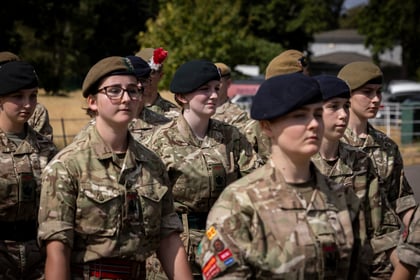 A group of young people in military uniforms and berets stand outdoors in formation, with trees and a building in the background. One person has a red flower in their beret.