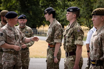 A group of soldiers in camouflage uniforms stand outdoors; an officer in the foreground appears to congratulate or address two young soldiers lined up in front of him. Trees and dry grass are in the background.