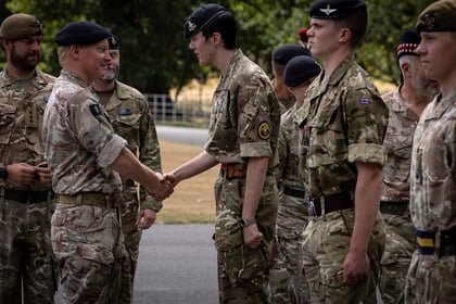 A military officer shakes hands with a young soldier in uniform while others in camouflage stand nearby outdoors, suggesting a ceremony or award event.