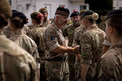 A group of soldiers in camouflage uniforms stand together outdoors; one man in a chequered beret speaks to a female soldier whilst others look on.
