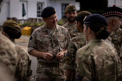 A group of uniformed military personnel gather outdoors, with one man in the centre talking and smiling to others. The scene appears informal and the background shows white buildings and greenery.