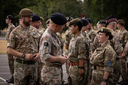A group of military personnel in camouflage uniforms and berets stand outdoors in formation, talking and smiling, with trees visible in the background.