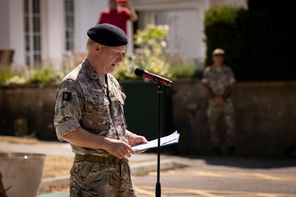 A man in military uniform and beret reads from papers at a microphone outdoors. Two other people in military attire are blurred in the background, with sunlight highlighting the scene.