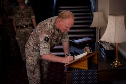 A person in military uniform signs a book on a desk next to a lamp, with other uniformed people standing in the background near a staircase.