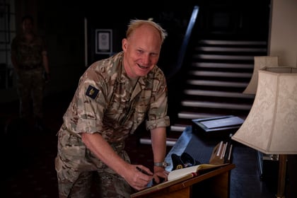 A smiling man in a camouflage military uniform is signing a book on a wooden stand indoors, with a table lamp beside him and stairs in the background.