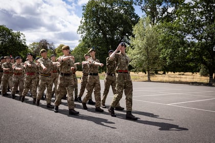 A group of people in camouflage uniforms march in formation on a paved area outdoors, surrounded by trees and greenery under a partly cloudy sky.
