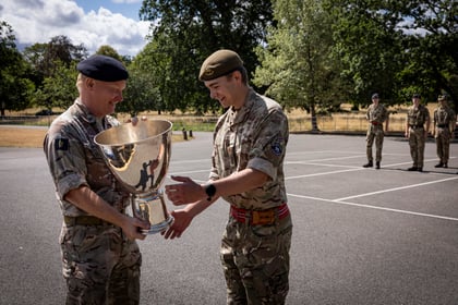 Two soldiers in camouflage uniforms and berets smile whilst holding a large trophy outdoors. Three other soldiers stand to attention in the background on a paved area with trees.