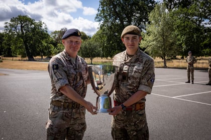 Two soldiers in camouflage uniforms hold a large silver trophy together outdoors. Trees and another uniformed person are visible in the background on a sunny day.