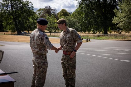 Two people in camouflage military uniforms shake hands outdoors in a paved open area, with trees and a grassy field in the background under a partly cloudy sky.