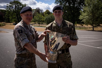 Two people in military uniforms stand outdoors, smiling and holding a large shiny trophy together. Trees and a clear sky are visible in the background.
