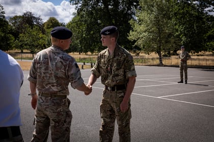 Two people in military uniforms shake hands outdoors on a paved area, with trees and a third person in uniform standing in the background. The weather is sunny with some clouds visible in the sky.