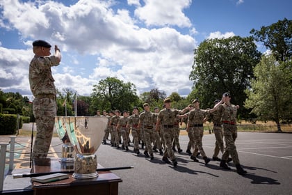 A group of soldiers in uniform march in formation outdoors while an officer stands on a platform, saluting them. Large silver trophies are displayed on a nearby table. Trees and clouds are visible in the background.