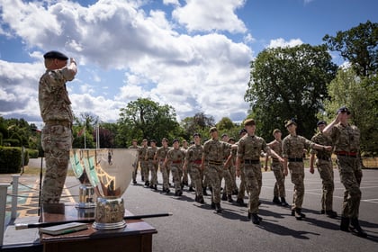 A group of uniformed cadets marches in formation outdoors, saluting a military officer standing beside silver trophies on a table. Trees and a partly cloudy sky are visible in the background.