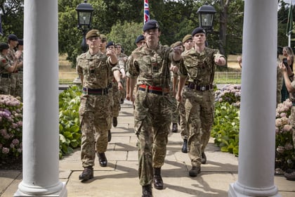 A group of soldiers in uniform march in formation under a white pergola, holding a Union Flag, with onlookers standing nearby and trees in the background.