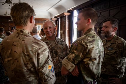 A group of soldiers in camouflage uniforms stand indoors, talking and smiling with each other. Sunlight filters through windows, casting warm light on the scene.