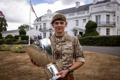A young person in military uniform smiles whilst holding a large silver trophy in front of a white building with manicured bushes and flags.