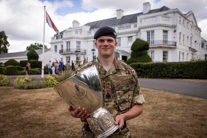 A young soldier in camouflage uniform and beret stands outside a large white building, smiling and holding a large silver trophy. A flag and several people are visible in the background.
