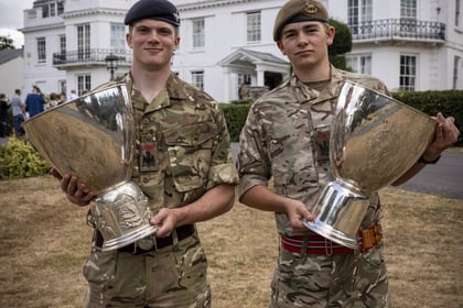 Two young soldiers in camouflage uniforms stand on grass, each holding a large silver trophy, with a white building and cloudy sky in the background.