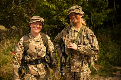 Two smiling Army Cadets in camouflage uniforms stand outdoors, one holding a rifle. Both have face paint and are surrounded by greenery under sunlight.