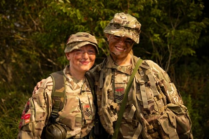 Two Army Cadets in camouflage uniforms with face paint stand outdoors, smiling and leaning towards each other. Green foliage is visible in the background.