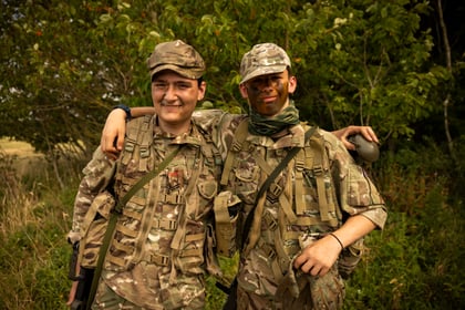 Two Army Cadets in camouflage uniforms and hats stand outdoors, smiling with arms round each other. One has face paint and holds a water bottle. Green foliage and tall grass surround them.