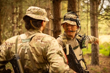 Two Army Cadets in camouflage kit stand in a forest. One is facing away, while the other points into the distance, giving directions. Both wear helmets and carry rifles. Sunlight filters through the trees.