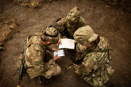 Three Army Cadets in camouflage uniforms kneel on the ground, gathered around open notebooks and a tablet, appearing to discuss or review plans or information outdoors.