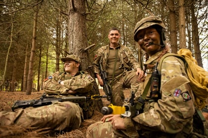 Three Army Cadets in camouflage uniforms sit and kneel in a forest. Two are smiling, one with camo face paint. They have military kit and rifles, surrounded by tall trees and dappled sunlight.