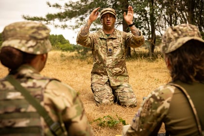 A soldier in camouflage uniform kneels on dry grass, gesturing with both hands while speaking to two Army Cadets seated in front of him, outdoors near trees.