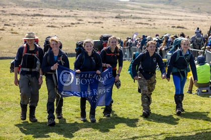 Six smiling hikers in outdoor gear walk on grass, holding a blue banner reading Sherborne Boys. They carry rucksacks and look happy, with a hilly landscape and more people in the background.