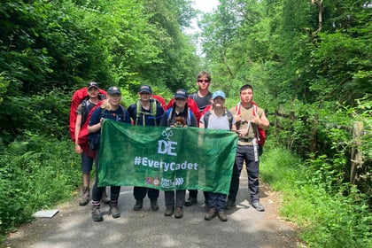 A group of cadets on a walking expedition stands in a gravel path holding a DofE flag