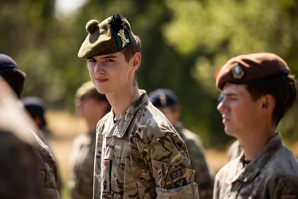 Young Army Cadets in military uniforms stand outdoors; one in focus wears a khaki beret with a feather, looking at the camera, whilst others in brown berets stand nearby. Sunlight and greenery are visible in the background.