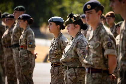 A group of young people in camouflage military uniforms stand in formation outdoors, focused and attentive, with trees and sunlight in the background.