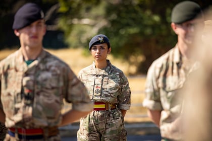 A female cadet in military uniform and beret stands in formation outdoors, looking ahead, with two male soldiers blurred in the foreground.