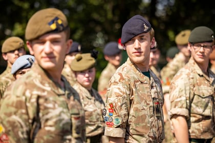 A group of Army Cadets in camouflage uniforms and various berets stand outdoors, some facing forwards and others to the side, in bright sunlight with trees blurred in the background.