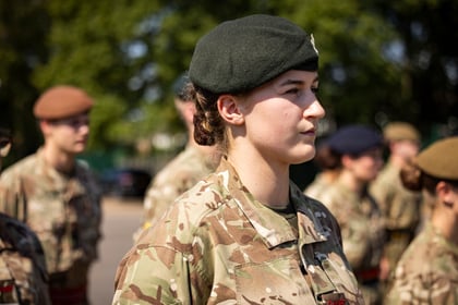 A group of Army Cadets in camouflage uniforms and berets stand outdoors in formation. The main focus is a woman in the foreground, looking ahead with a serious expression. Trees and more soldiers are blurred in the background.