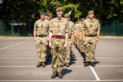 A group of Army Cadets in camouflage uniforms stand in formation outdoors on a sunny day, with trees and a fence in the background. The front cadet appears to be giving a command.