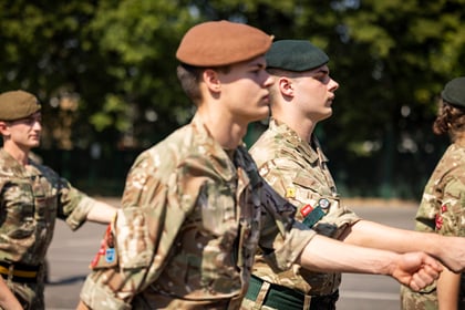 Several Army Cadets in camouflage uniforms and berets march in formation outdoors on a sunny day, with green trees in the background.