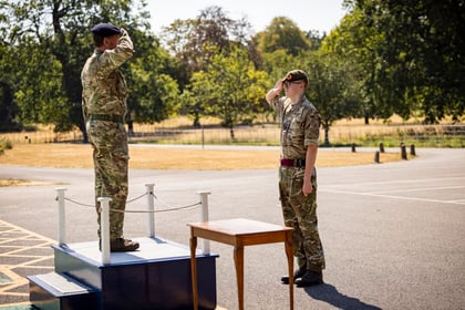 An Army Cadet salutes an officer outdoors, one standing on a platform and the other beside a wooden table, with trees and grass visible in the background.