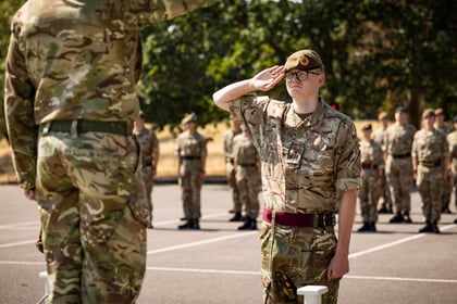 A group of Army Cadets in camouflage uniforms stand in formation outdoors. One cadet in the foreground salutes another officer, with trees visible in the background.