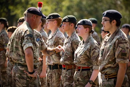 A group of uniformed Army Cadets stands in a row outdoors, talking to a higher-ranking officer. They wear camo uniforms and berets, with trees visible in the background.