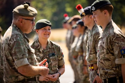 A group of people in Army Cadet uniforms stand outside in sunlight, talking and smiling. The focus is on two individuals in conversation, with others in the background.