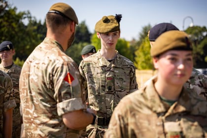 A group of Army Cadets in camouflage uniforms and berets stand outdoors in sunlight, some interacting and smiling, with trees and clear sky in the background.