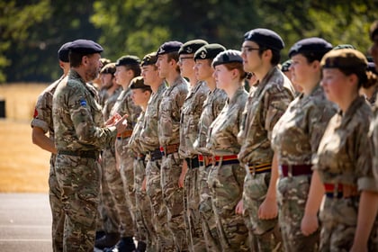 A group of uniformed cadets stand in formation outdoors as an instructor in similar attire addresses them, with trees blurred in the background.